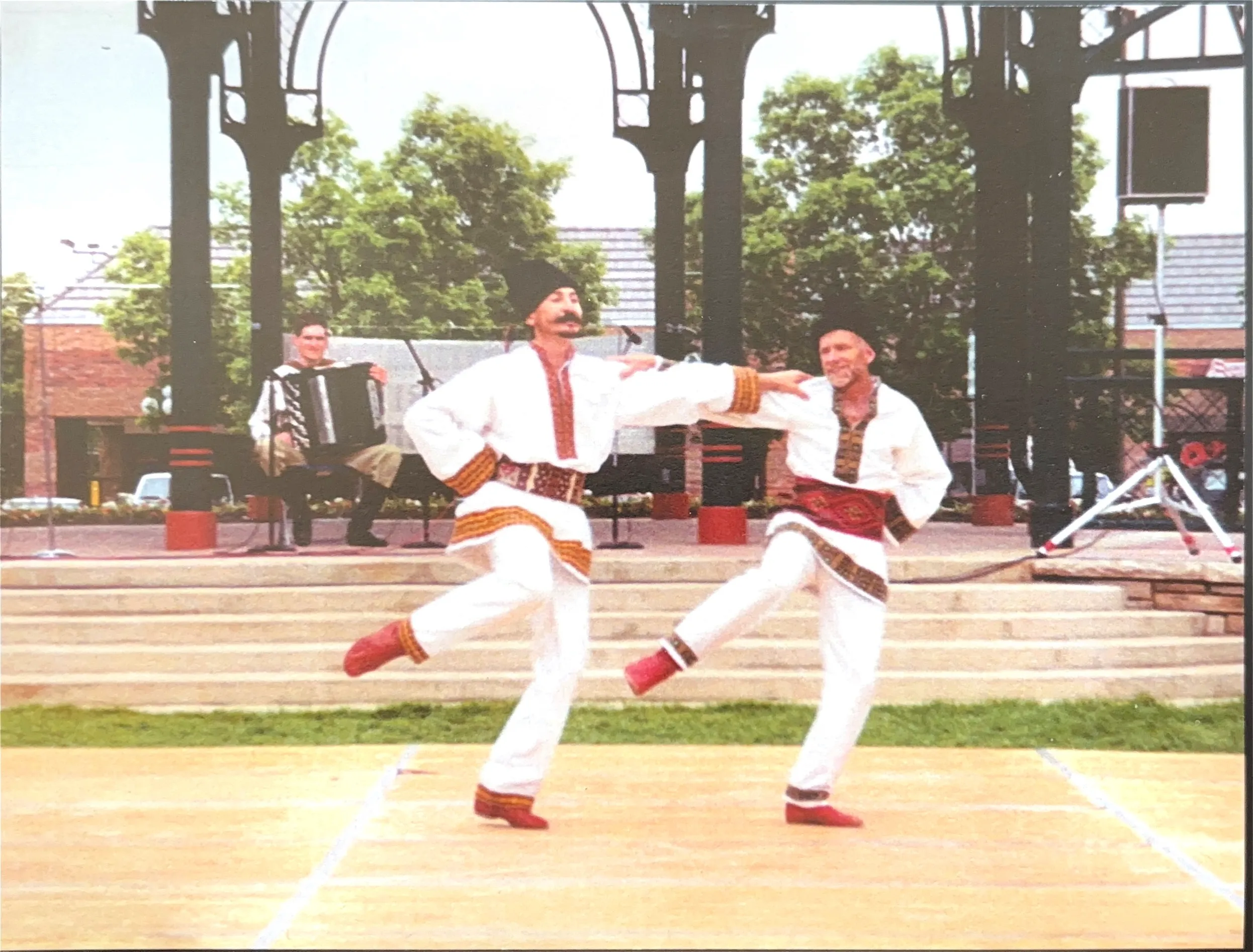 Two pairs of postoley folk dancers dancing on a stage in a park.